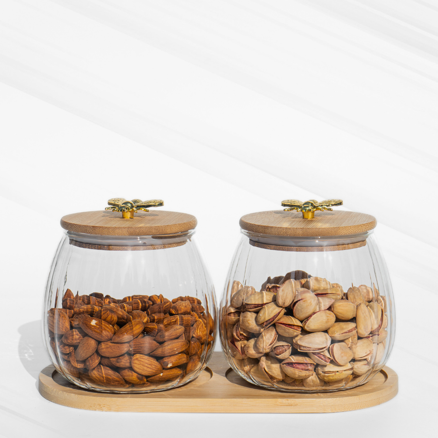 A set of two fluted borosilicate glass storage jars with airtight wooden lids featuring gold butterfly knobs, placed on a matching wooden tray for elegant storage.