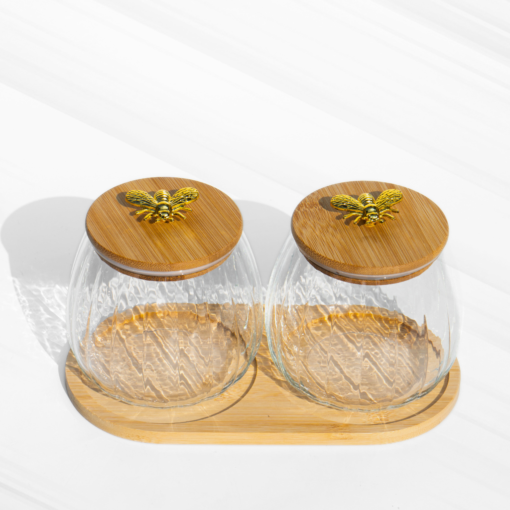 A set of two fluted borosilicate glass storage jars with airtight wooden lids featuring gold butterfly knobs, placed on a matching wooden tray for elegant storage.