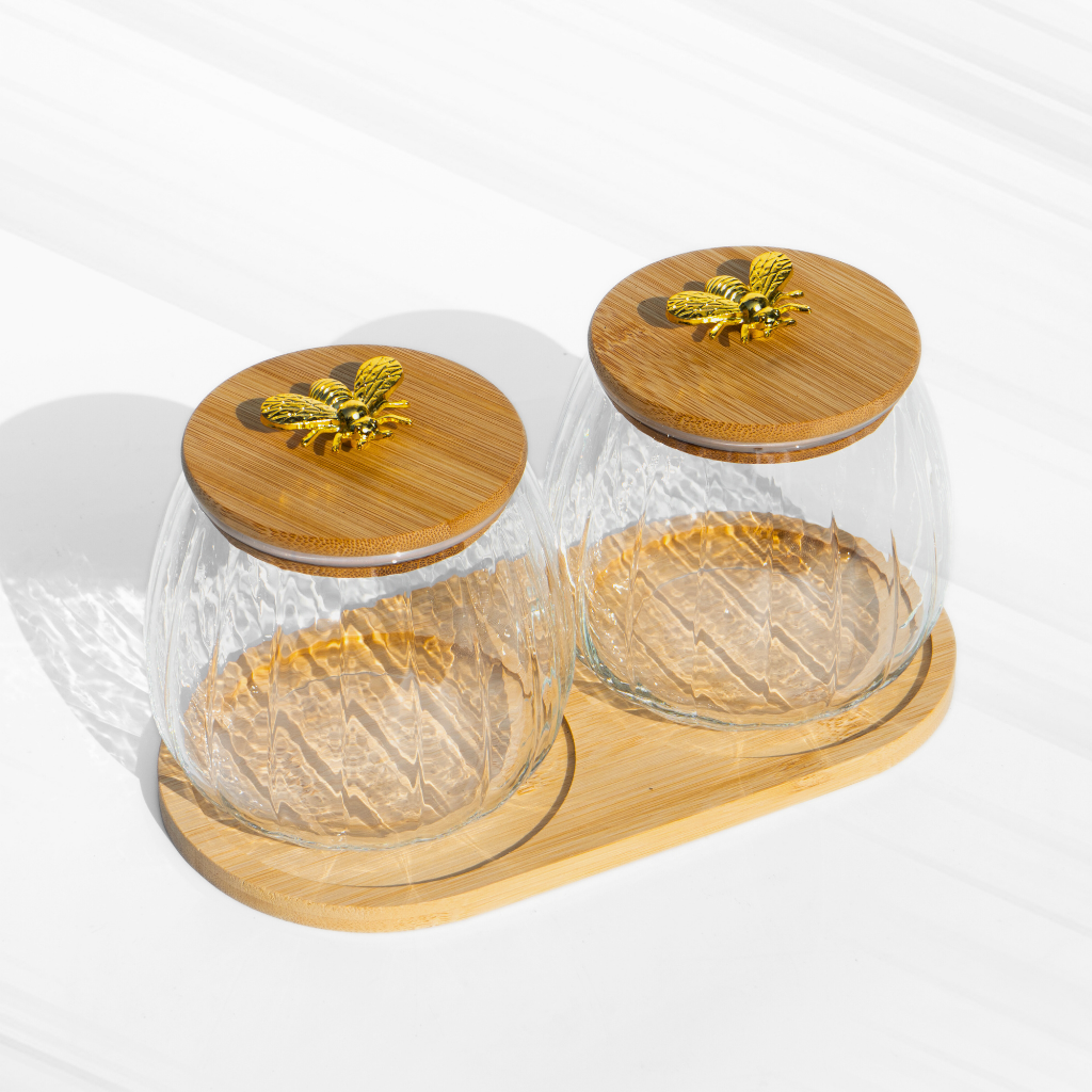 A set of two fluted borosilicate glass storage jars with airtight wooden lids featuring gold butterfly knobs, placed on a matching wooden tray for elegant storage.