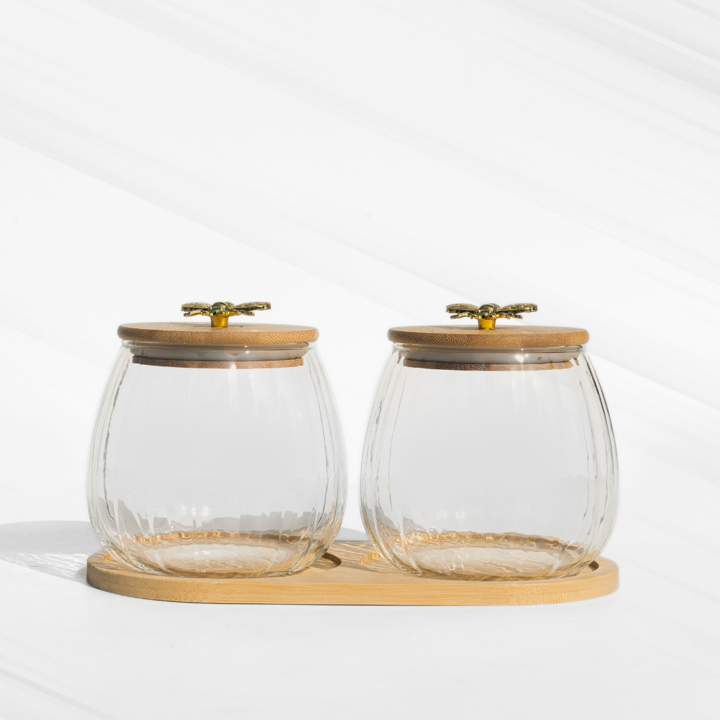 A set of two fluted borosilicate glass storage jars with airtight wooden lids featuring gold butterfly knobs, placed on a matching wooden tray for elegant storage.